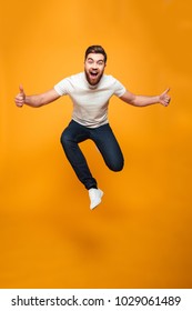 Full length portrait of an excited bearded man jumping and showing thumbs up isolated over yellow background