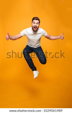 Full length portrait of an excited bearded man jumping and showing thumbs up isolated over yellow background