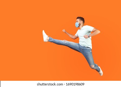 Full length portrait of excited man with surgical medical mask in casual style jumping over in air, running quickly fast, in hurry for discounts. indoor studio shot isolated on orange background