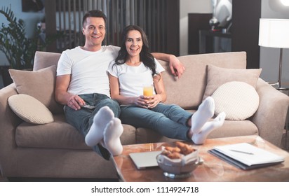 Full Length Portrait Of Excited Married Couple Is Watching TV With Interest. They Are Hugging While Sitting In Living Room. Lovers Are Putting Legs On Small Table 