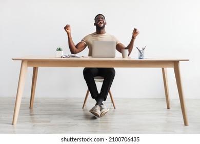 Full length portrait of excited millennial black man gesturing YES in front of laptop, celebrating success or achievement, sitting at desk in home office, copy space. Remote job, online win concept