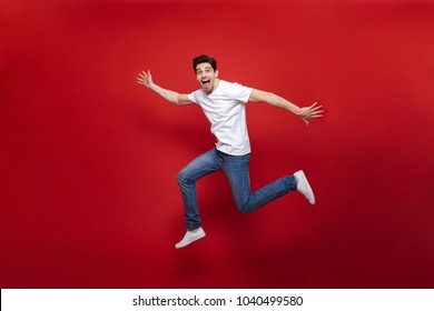 Full Length Portrait Of An Excited Young Man In White T-shirt Jumping While Celebrating Success Isolated Over Red Background