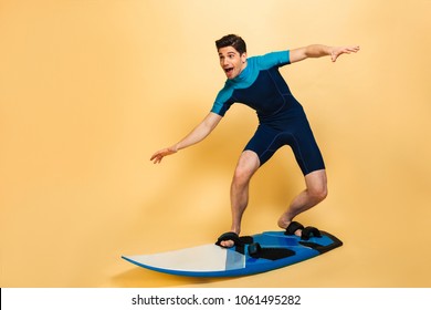 Full length portrait of an excited young man dressed in swimsuit surfing on a board isolated over yellow background - Powered by Shutterstock