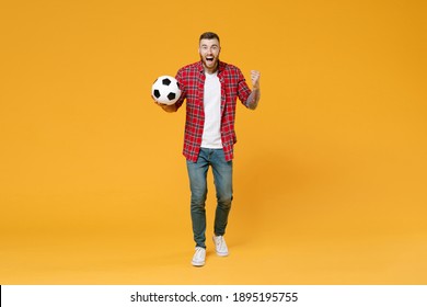 Full length portrait of excited young man football fan in red shirt cheer up support favorite team with soccer ball clenching fists isolated on yellow background studio. People sport leisure concept