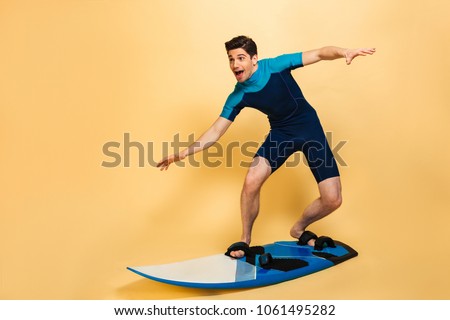 Full length portrait of an excited young man dressed in swimsuit surfing on a board isolated over yellow background