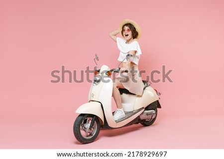 Full length portrait of excited young brunette woman wearing white summer clothes hat glasses keeping mouth open looking aside sitting driving moped isolated on pastel pink colour background studio