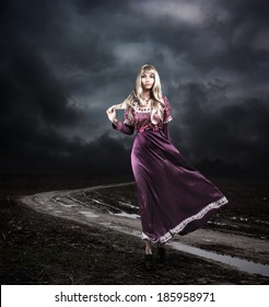 Full Length Portrait Of Fantasy Woman In Waving Purple Dress Walking On Dirty Road. Moody Sky. HDR Cloudscape.