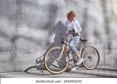 Full Length Portrait Of Fashionable Woman In Business Casual Clothes Holding Her Hands On Handlebar Of A City Bike Isolated On The Grey Wall