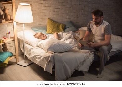 Full Length Portrait Of Father Reading Bedtime Story To Son At Night Sitting On Edge Of Bed In Dim Lighting, Copy Space