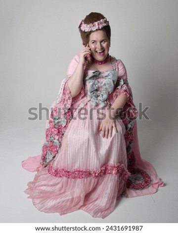 Full length portrait female model wearing opulent pink gown costume of historical French baroque nobility, style of Marie Antoinette. Sitting pose on throne using modern mobile phone technology.
