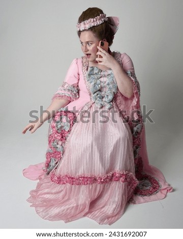 Full length portrait female model wearing opulent pink gown costume of historical French baroque nobility, style of Marie Antoinette. Sitting pose on throne using modern mobile phone technology.
