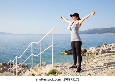 Full Length Portrait Of Fit Mature Woman Standing With Arms Raised By The Sea After Workout While Relaxing. 