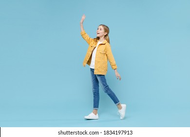 Full length portrait of funny little blonde kid girl 12-13 years old in yellow jacket isolated on blue background studio. Childhood lifestyle concept. Waving and greeting with hand as notices someone