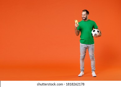 Full length portrait funny man football fan in basic green t-shirt cheer up support favorite team with soccer ball using mobile cell phone isolated on orange background. People sport leisure concept