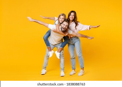 Full Length Portrait Funny Parents Mom Dad With Child Kid Daughter Teen Girl In T-shirts Giving Piggyback Ride To Joyful Sit On Back Spreading Hands Isolated On Yellow Background. Family Day Concept