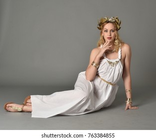 Full Length Portrait Of Girl Wearing White Ancient Greek Or Roman Costume, Seated
 Pose On A Grey Background.