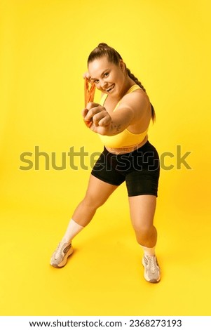 Full length portrait of grimacing plus size sports woman in tracksuit training hard with sport elastic bands for hands over yellow background. Concept of sport, hobby, health, lifestyle, workout