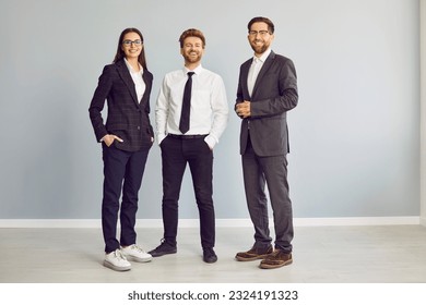 Full length portrait of a group of three young cheerful business people in suits standing confidently, looking at the camera and smiling isolated on a studio grey background. Team work concept. - Powered by Shutterstock