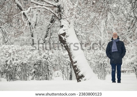 Full length portrait of handome mature man in trendy winter clothing and fashionable glasses standing in snow park. Text space. Outdoor shot