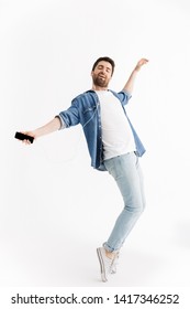 Full Length Portrait Of A Handsome Bearded Man Wearing Casual Clothes Jumping Isolated Over White Background, Listening To Music With Earphones, Holding Mobile Phone, Dancing