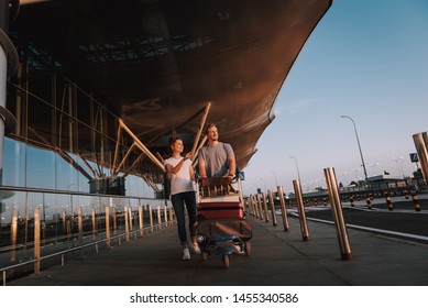 Full length portrait of handsome bearded man pushing luggage trolley and looking away with smile while his charming girlfriend pointing at something interesting