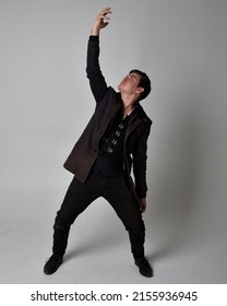 Full length portrait of  handsome brunette male model wearing black leather coat and elegant vest. Standing Pose with gestural hands reaching out,  isolated on studio background.