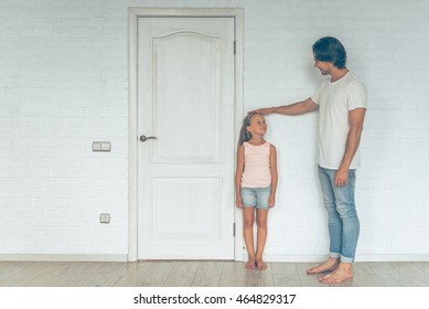 Full Length Portrait Of Handsome Father Measuring Height Of His Cute Little Daughter Near The Door At Home. Both Are Looking At Each Other And Smiling