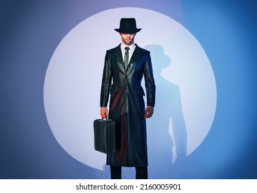 Full length portrait of handsome man in black coat and hat holding briefcase posing in the spotlight on studio background. noir film style. Private detective, spy, investigation concept. 