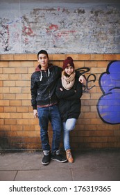 Full Length Portrait Of Handsome Young Man Standing With His Beautiful Girlfriend Leaning Against A Wall. Mixed Race Teenage Couple Posing Together Outdoors