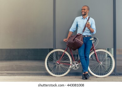 Full length portrait of handsome young Afro American man in casual clothes using phone, looking away and smiling while leaning on his bike, standing outdoors