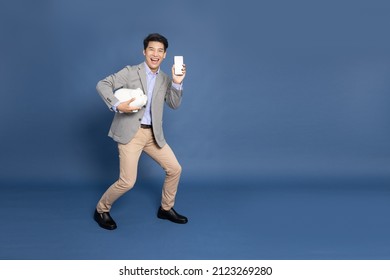 Full Length Portrait Of Happy Asian Businessman Holding White Piggy Bank And Mobile Phone Isolated On Blue Background, Business And Financial Concept