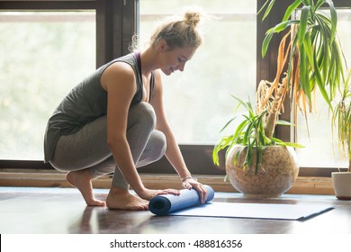 Full Length Portrait Of Happy Attractive Young Woman Folding Blue Yoga Or Fitness Mat After Working Out At Home In Living Room. Healthy Life, Keep Fit Concepts. Horizontal Image