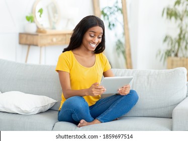 Full length portrait of happy black woman using tablet computer on couch in living room. African American lady with touch pad browsing web, watching video or communicating on webcam - Shutterstock ID 1859697514