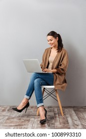 Full length portrait of a happy casual asian woman holding laptop computer while sitting on a chair over gray background