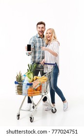 Full length portrait of a happy couple showing blank screen mobile phone and pointing finger while standing with a supermarket trolley isolated over white background