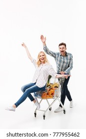 Full Length Portrait Of A Happy Couple Having Fun With A Supermarket Trolley And Waving Isolated Over White Background