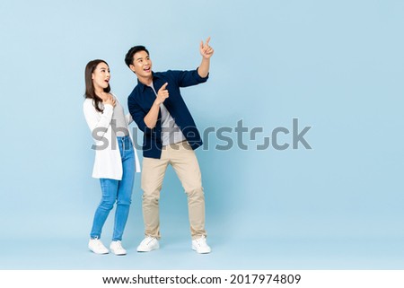 Full length portrait of happy excited Asian couple tourists pointing hands to empty space on isolated light blue background