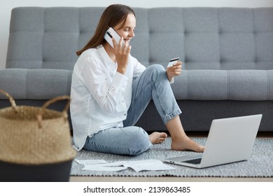 Full length portrait of happy exited woman wearing white shirt and jeans sitting on floor near sofa, using laptop and talking phone, holding credit card and expressing happiness.
