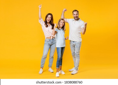 Full Length Portrait Of Happy Joyful Funny Young Parents Mom Dad With Child Kid Daughter Teen Girl In T-shirts Clenching Fists Doing Winner Gesture Isolated On Yellow Background. Family Day Concept