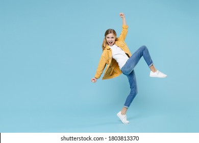 Full Length Portrait Of Happy Little Blonde Kid Girl 12-13 Years Old In Yellow Jacket Clenching Fists Like Winner, Say Yeah, Raised Up Leg Isolated On Blue Wall Background. Childhood Lifestyle Concept