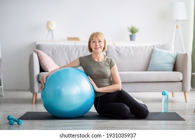 Full length portrait of happy mature woman resting on yoga mat with fitness ball, smiling at camera indoors, free space. Cheerful senior lady taking break from her domestic training