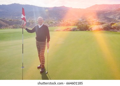 Full Length Portrait Of Happy Senior Male Golfer Holding Flag And Putter At Golf Course