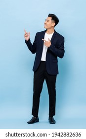 Full length portrait of happy smiling young handsome southeast Asian businessman pointing and looking upward on light blue studio background