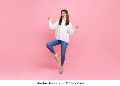 Full length portrait of happy woman doing victory gesture, showing peace, v sign with double fingers, wearing white casual style sweater. Indoor studio shot isolated on pink background.