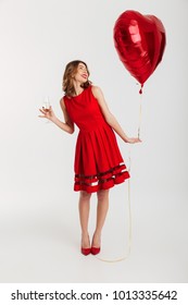 Full Length Portrait Of A Happy Young Woman Dressed In Red Dress Holding Air Balloon While Drinking Glass Of Champagne And Celebrating Isolated Over White Background