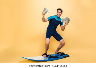 Full length portrait of a happy young man dressed in swimsuit holding bunch of money banknotes while surfing on a board isolated over yellow background - Powered by Shutterstock