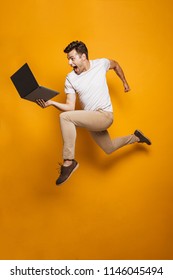 Full Length Portrait Of A Happy Young Man Jumping With Laptop Computer Isolated Over Yellow Background