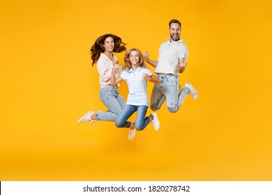 Full Length Portrait Of Happy Young Parents Mom Dad With Child Kid Daughter Teen Girl In Basic T-shirts Jumping Doing Winner Gesture Isolated On Yellow Background Studio Portrait. Family Day Concept