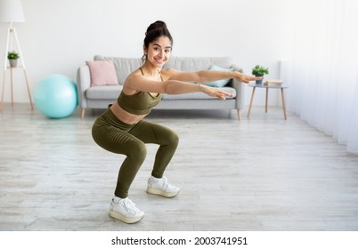 Full length portrait of happy young Indian woman doing squats exercises at home, blank space. Cheerful millennial Asian lady in sportswear performing strength exercises, keeping in great shape