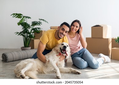 Full Length Portrait Of Happy Young Diverse Couple With Their Dog Posing On Floor Of New Home On Moving Day. Millennial Homeowners With Cute Pet Sitting Among Cardboard Boxes, Relocating To New House